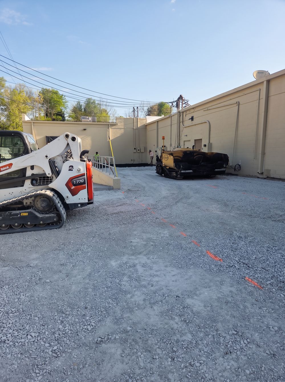 Skid steer and asphalt paver parked on a stone base beside a building